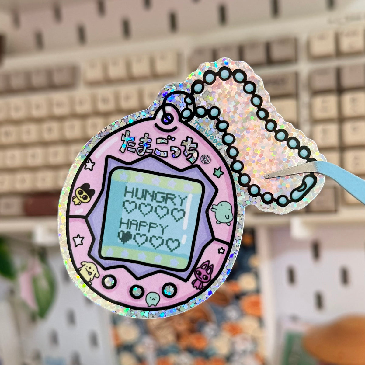 Tama Hungry Glitter Sticker featuring pink and purple virtual pet device with Hungry Hungry lettering.