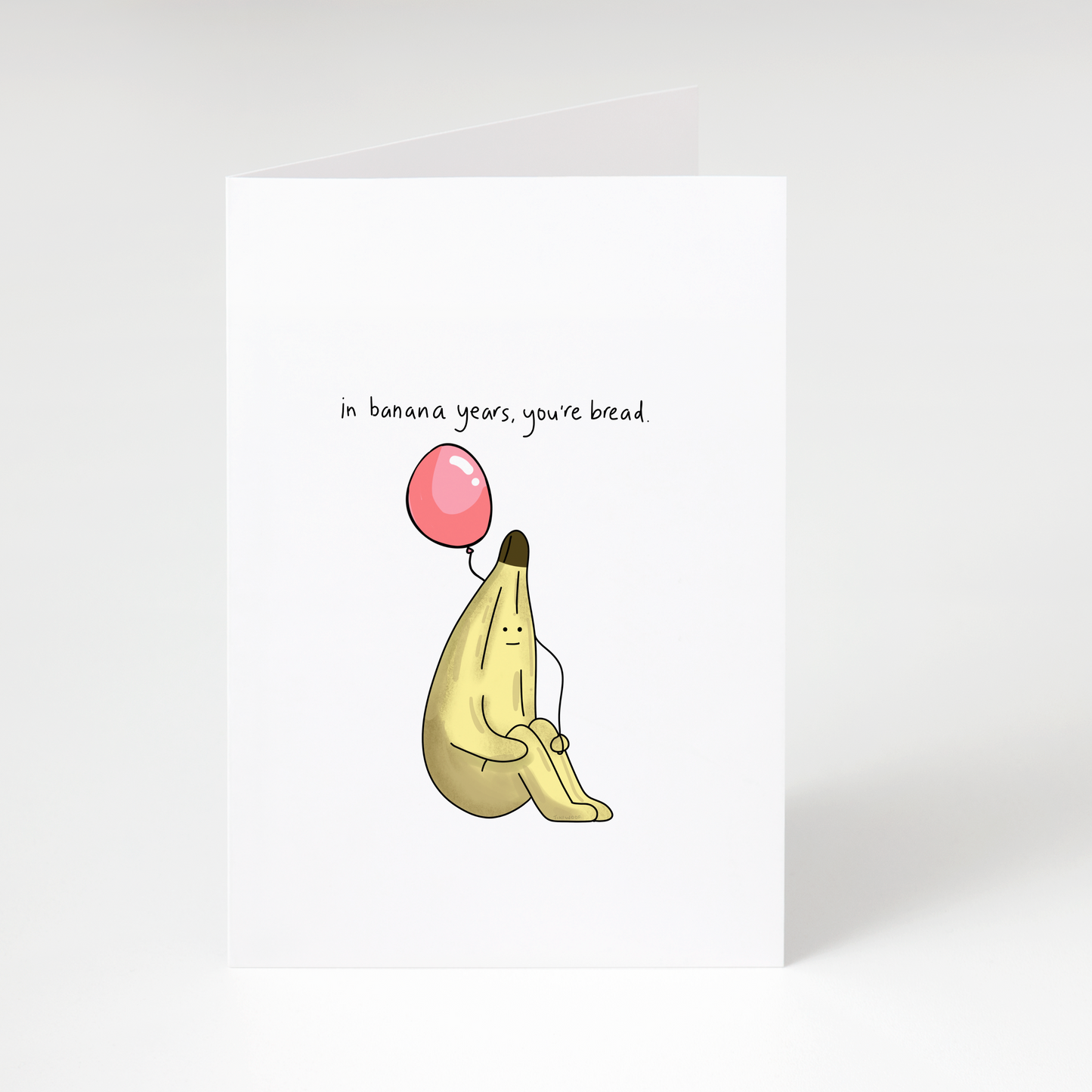 Cute banana holding a pink balloon with text “In banana years, you’re bread” on white birthday greeting card