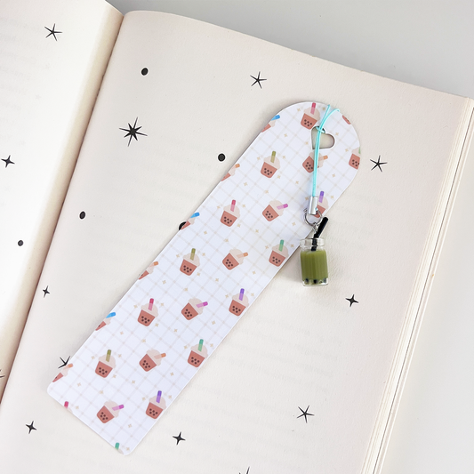 White grid-patterned bookmark with cute bubble tea illustrations and a mini green matcha boba drink charm on an open book.
