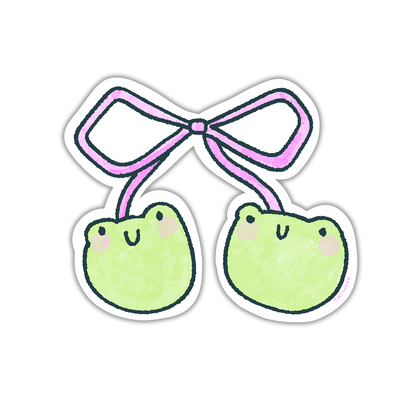 Hand holding a cute vinyl sticker of two pastel green frog faces tied together with a pink bow, illustrated in a kawaii Tiki Woof Studio style.
