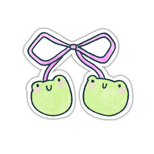 Hand holding a cute vinyl sticker of two pastel green frog faces tied together with a pink bow, illustrated in a kawaii Tiki Woof Studio style.