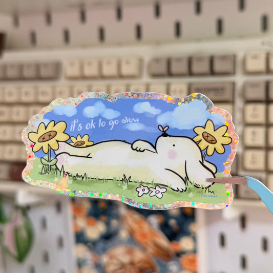 It's Okay to Go Slow Glitter Sticker featuring sleeping white animal among daisies below a blue sky.
