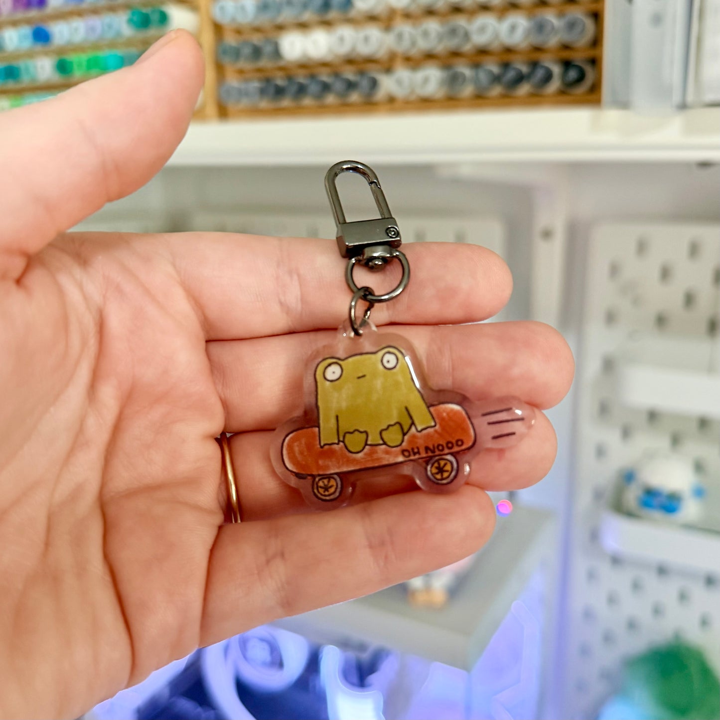 Hand holding a keychain with a cartoon character design in front of a pegboard background.