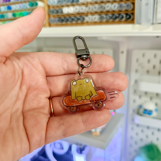 Hand holding a keychain with a cartoon character design in front of a pegboard background.