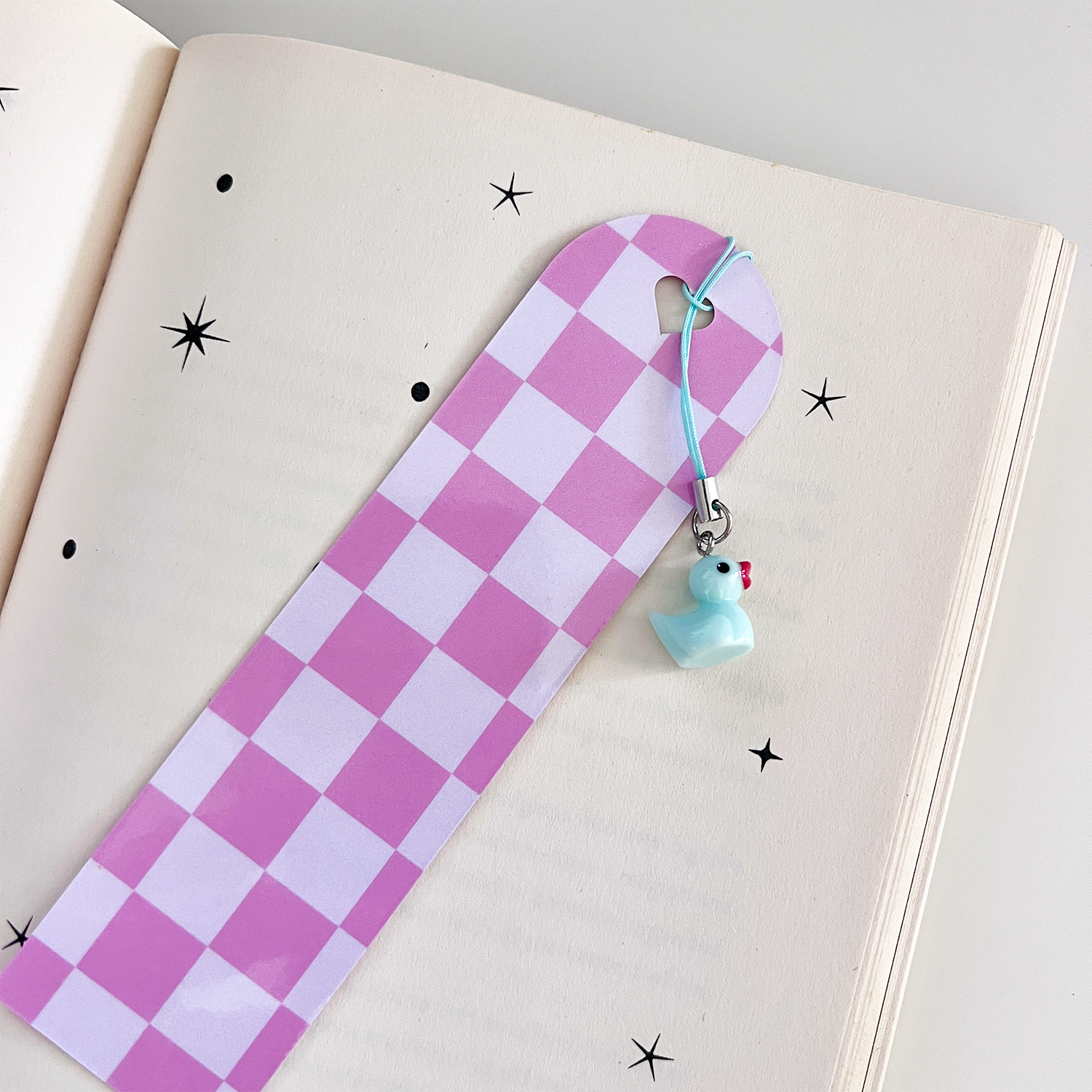 Pink checkered bookmark with a pastel blue duck charm resting on an open book with star doodles.
