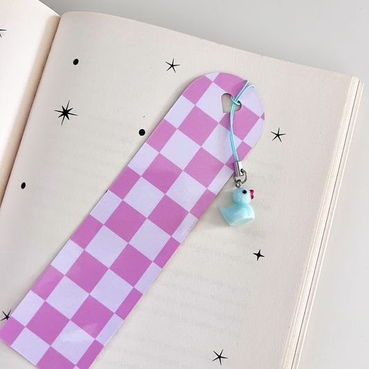 Pink checkered bookmark with a pastel blue duck charm resting on an open book with star doodles.