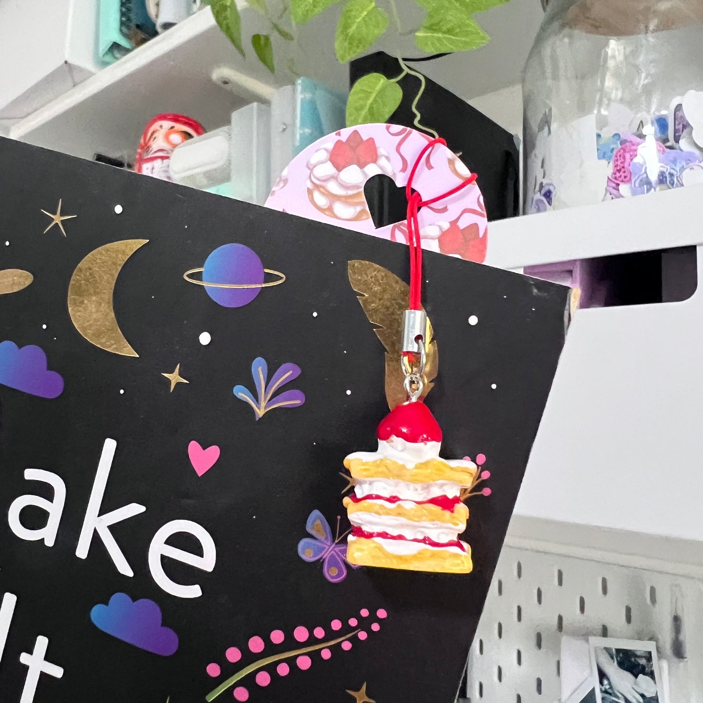 A pastel purple bookmark decorated with repeating strawberry cake illustrations lies open on a book page. A matching miniature strawberry cake charm hangs from a red cord at the top of the bookmark.