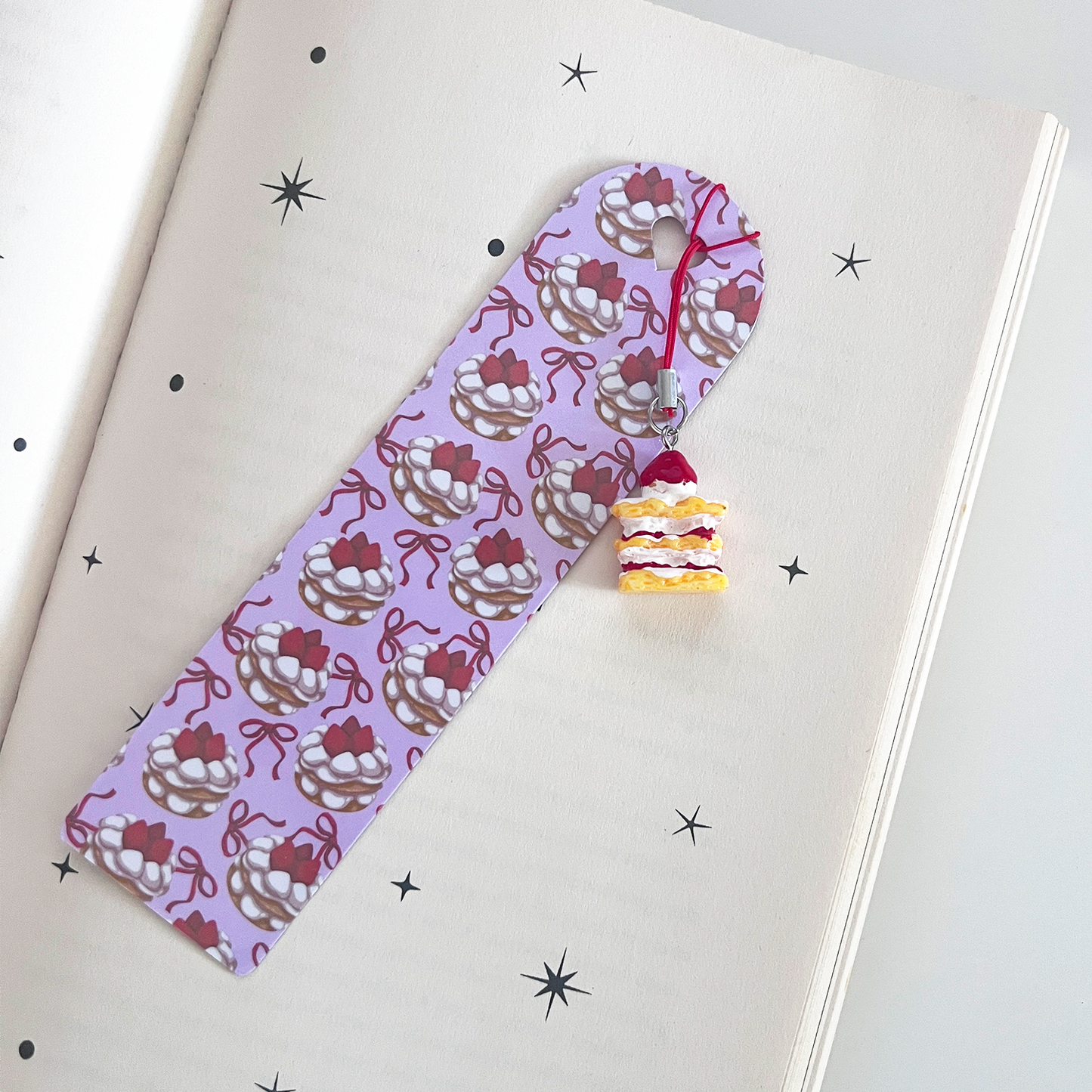 A pastel strawberry cake themed bookmark is clipped over the top of a book cover. A small strawberry cake charm with red cord hangs from the bookmark, displayed against a celestial-themed book cover.