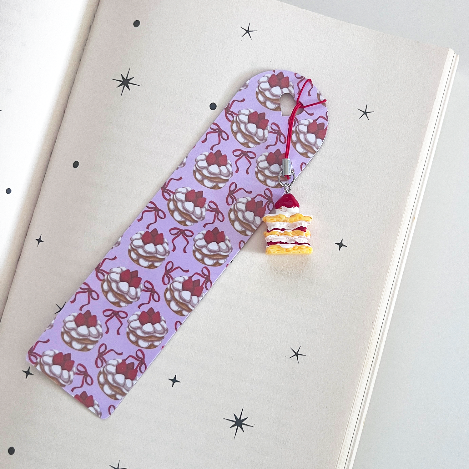 A pastel strawberry cake themed bookmark is clipped over the top of a book cover. A small strawberry cake charm with red cord hangs from the bookmark, displayed against a celestial-themed book cover.