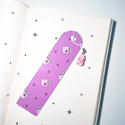 A purple kawaii bookmark with small strawberry milk carton illustrations lies open on a book. A pink strawberry milk charm hangs from the top on a white cord, creating a cute pastel aesthetic.
