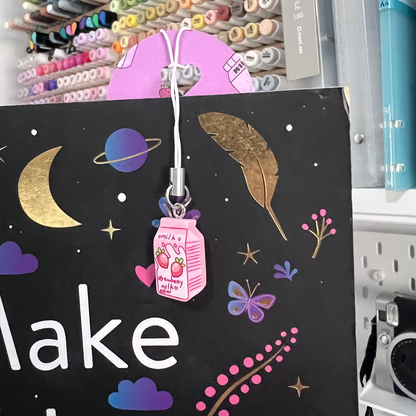 A close-up of the strawberry milk kawaii bookmark hanging over the top of a book. The pink milk-carton charm dangles in front of a dark cover decorated with moons, planets, stars, and gold accents.